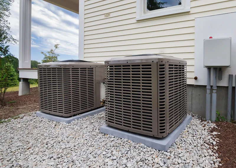 Two outdoor residential HVAC condenser units on gravel pads next to a beige-sided house