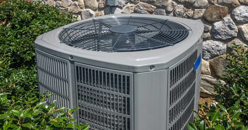 Outdoor HVAC condenser unit beside stone wall, surrounded by green shrubs