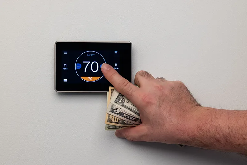 A hand adjusts a smart thermostat to 70°F while holding U.S. dollar bills, illustrating energy cost savings.