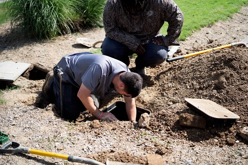Two workers repairing an underground irrigation valve in a dirt yard, one man reaching into a meter box with shovels nearby