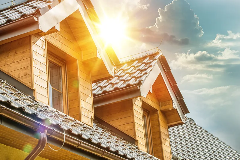 Sunlit wooden dormer windows on a tiled roof with sun flare and partly cloudy sky